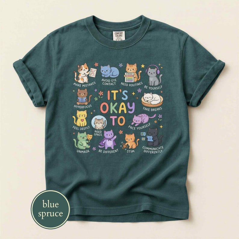May include: A dark teal short-sleeve t-shirt featuring a graphic of various cartoon cats with the text "IT'S OKAY TO" surrounded by phrases like "AVOID EYE CONTACT," "BE YOURSELF," "HYPERFOCUS," "FEEL DEEPLY," "NEED SPACE," "PACE," "STIM," and "COMMUNICATE DIFFERENTLY."