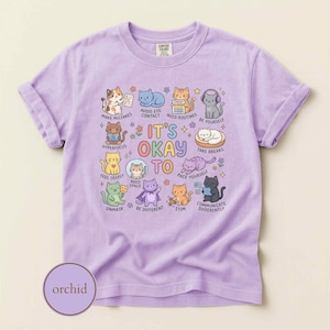 May include: A light purple t-shirt featuring a graphic print of various cartoon cats with the text IT'S OKAY TO, MAKE MISTAKES, AVOID EYE CONTACT, NEED ROUTINES, BE YOURSELF, HYPERFOCUS, FEEL DEEPLY, NEED SPACE, TAKE BREAKS, UNMASK, BE DIFFERENT, STIM, COMMUNICATE DIFFERENTLY.