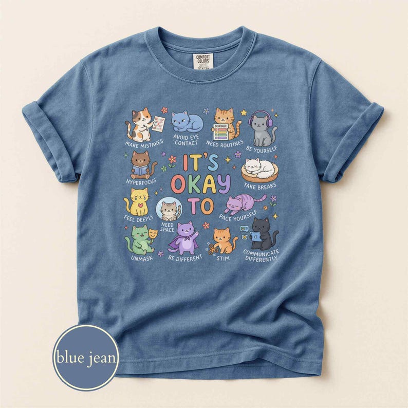 May include: A blue jean colored t-shirt featuring a graphic print of various cartoon cats with the text IT'S OKAY TO and phrases including NEED SPACE, UNMASK, BE DIFFERENT, STIM, COMMUNICATE DIFFERENTLY, TAKE BREAKS, and HYPERFOCUS.
