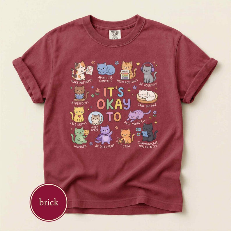 May include: A brick red short-sleeve t-shirt featuring a graphic print of various cartoon cats with the text IT'S OKAY TO and labels including MAKE MISTAKES, AVOID EYE CONTACT, NEED ROUTINES, BE YOURSELF, HYPERFOCUS, TAKE BREAKS, FEEL DEEPLY, NEED SPACE, PACE YOURSELF, UNMASK, BE DIFFERENT, STIM, and COMMUNICATE DIFFERENTLY.