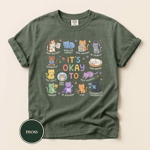 May include: A moss green t-shirt featuring a graphic of eleven cartoon cats with the text IT'S OKAY TO and various phrases including MAKE MISTAKES, AVOID EYE CONTACT, NEED ROUTINES, BE YOURSELF, HYPERFOCUS, TAKE BREAKS, FEEL DEEPLY, NEED SPACE, UNMASK, BE DIFFERENT, STIM, and COMMUNICATE DIFFERENTLY.