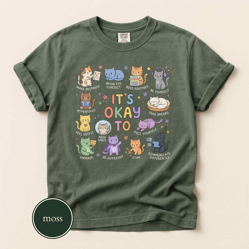 May include: A moss green t-shirt featuring a graphic of eleven cartoon cats with the text IT'S OKAY TO and various phrases including MAKE MISTAKES, AVOID EYE CONTACT, NEED ROUTINES, BE YOURSELF, HYPERFOCUS, TAKE BREAKS, FEEL DEEPLY, NEED SPACE, UNMASK, BE DIFFERENT, STIM, and COMMUNICATE DIFFERENTLY.