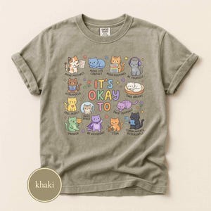 May include: A khaki crew neck t-shirt featuring a central graphic of various colorful cartoon cats with the text IT'S OKAY TO and labels including MAKE MISTAKES, AVOID EYE CONTACT, NEED ROUTINES, BE YOURSELF, HYPERFOCUS, FEEL DEEPLY, NEED SPACE, TAKE BREAKS, UNMASK, BE DIFFERENT, STIM, and COMMUNICATE DIFFERENTLY.