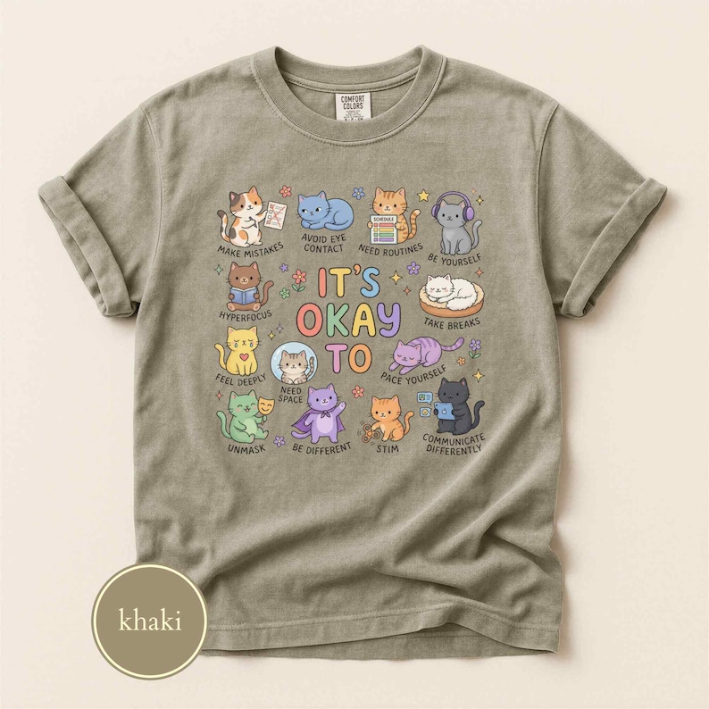 May include: A khaki crew neck t-shirt featuring a central graphic of various colorful cartoon cats with the text IT'S OKAY TO and labels including MAKE MISTAKES, AVOID EYE CONTACT, NEED ROUTINES, BE YOURSELF, HYPERFOCUS, FEEL DEEPLY, NEED SPACE, TAKE BREAKS, UNMASK, BE DIFFERENT, STIM, and COMMUNICATE DIFFERENTLY.