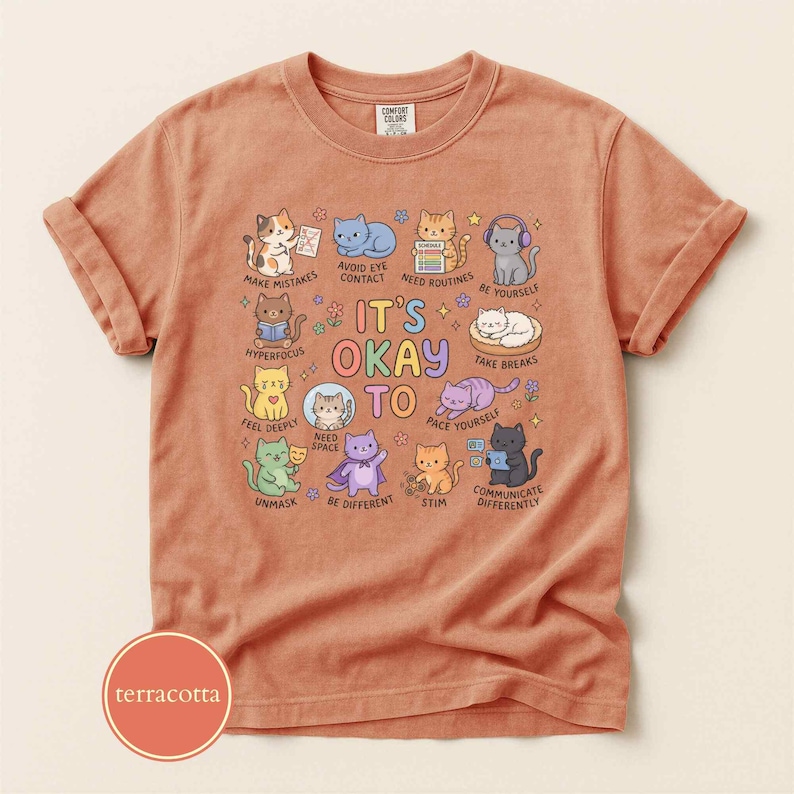 May include: A terracotta-colored t-shirt featuring a graphic print of various cartoon cats with the text "IT'S OKAY TO" surrounded by phrases like "MAKE MISTAKES," "NEED SPACE," "STIM," "UNMASK," "BE DIFFERENT," and "COMMUNICATE DIFFERENTLY."