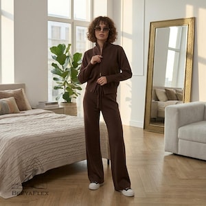 May include: A brown knitwear set consisting of a zip-up jacket and wide-leg pants. The jacket has a high collar and a drawstring waist. The pants have a drawstring waist. The model is wearing white sneakers. The set is displayed in a well-lit room with a large mirror.