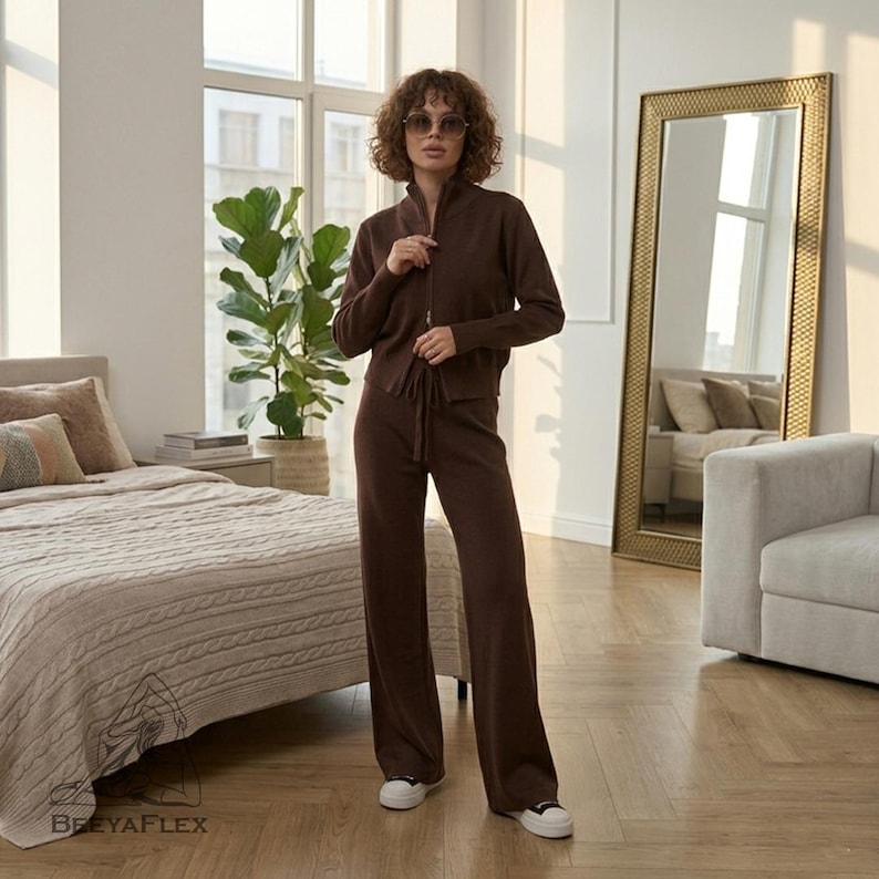 May include: A brown knitwear set consisting of a zip-up jacket and wide-leg pants. The jacket has a high collar and a drawstring waist. The pants have a drawstring waist. The model is wearing white sneakers. The set is displayed in a well-lit room with a large mirror.
