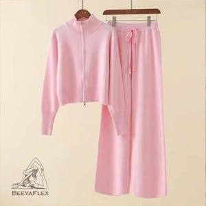 May include: A matching pink knitwear set, including a zip-up sweater with a high collar and wide-leg pants with a drawstring. The set is displayed on a wooden hanger. The sweater has long sleeves and a ribbed hem. The pants have a relaxed fit.