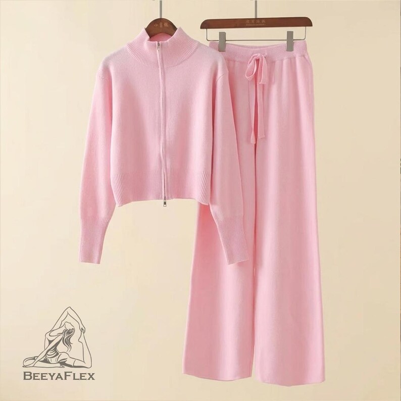 May include: A matching pink knitwear set, including a zip-up sweater with a high collar and wide-leg pants with a drawstring. The set is displayed on a wooden hanger. The sweater has long sleeves and a ribbed hem. The pants have a relaxed fit.