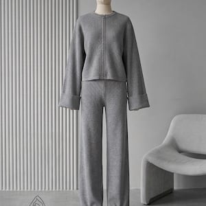 Puede incluir: Un conjunto de punto gris sobre un maniqu&iacute;. El conjunto incluye una chaqueta con cremallera y mangas anchas y pantalones anchos a juego. El atuendo es de color gris liso, y el maniqu&iacute; tiene un tono de piel neutro.