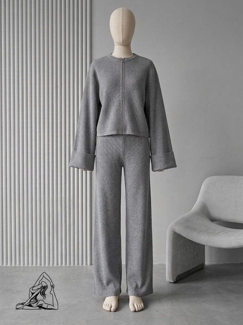 Puede incluir: Un conjunto de punto gris sobre un maniqu&iacute;. El conjunto incluye una chaqueta con cremallera y mangas anchas y pantalones anchos a juego. El atuendo es de color gris liso, y el maniqu&iacute; tiene un tono de piel neutro.
