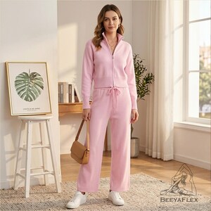 May include: A light pink knit tracksuit featuring a zip-up jacket with a high collar and matching drawstring pants. The outfit is paired with white sneakers and a small brown purse. The ensemble is displayed in a well-lit room with a framed botanical print.