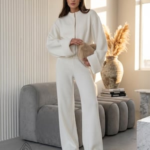 Puede incluir: Un conjunto de sal&oacute;n crema de dos piezas con una chaqueta con cremallera y pantalones de pernera ancha. El atuendo se combina con zapatos de plataforma color beige y un bolso de mano esponjoso marr&oacute;n claro. El conjunto se exhibe en una habitaci&oacute;n bien iluminada con un sof&aacute; gris y un jarr&oacute;n decorativo.