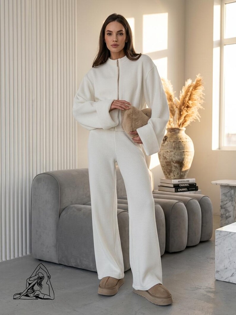 Puede incluir: Un conjunto de sal&oacute;n crema de dos piezas con una chaqueta con cremallera y pantalones de pernera ancha. El atuendo se combina con zapatos de plataforma color beige y un bolso de mano esponjoso marr&oacute;n claro. El conjunto se exhibe en una habitaci&oacute;n bien iluminada con un sof&aacute; gris y un jarr&oacute;n decorativo.