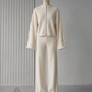Puede incluir: Conjunto de punto color crema, que incluye una chaqueta con cremallera y mangas anchas y pantalones de pernera ancha a juego. El conjunto se exhibe en un maniqu&iacute; sobre un fondo neutro, mostrando una est&eacute;tica minimalista. La tela parece suave y c&oacute;moda.