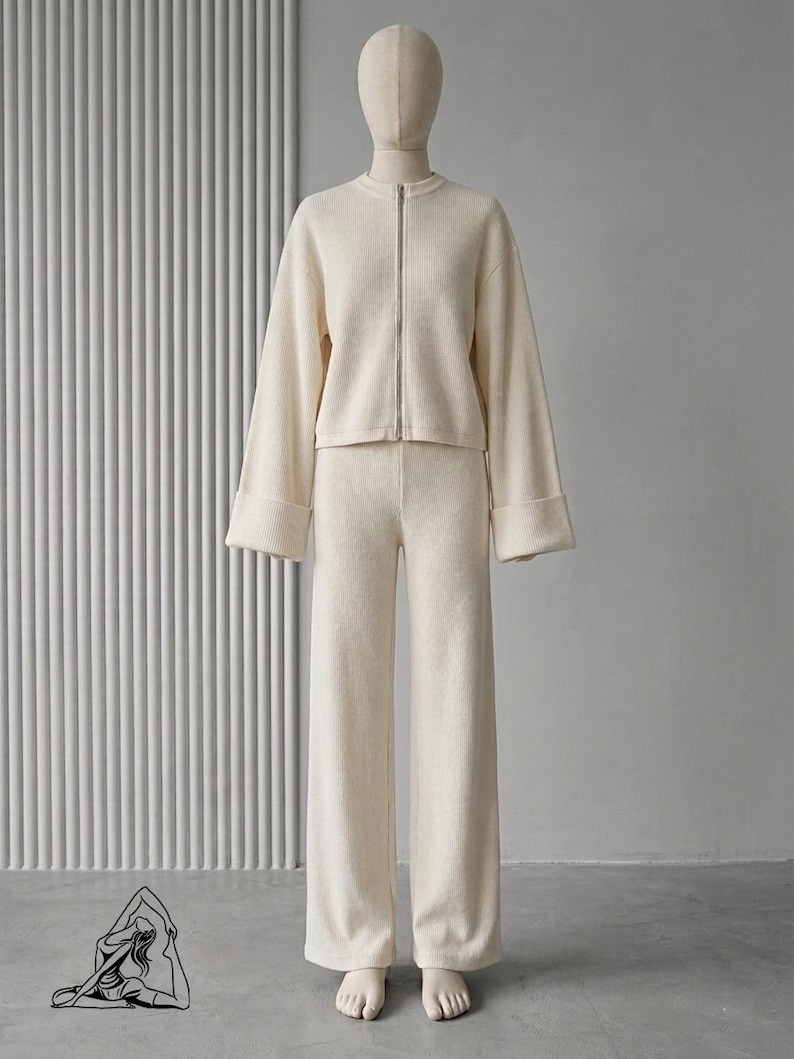 Puede incluir: Conjunto de punto color crema, que incluye una chaqueta con cremallera y mangas anchas y pantalones de pernera ancha a juego. El conjunto se exhibe en un maniqu&iacute; sobre un fondo neutro, mostrando una est&eacute;tica minimalista. La tela parece suave y c&oacute;moda.