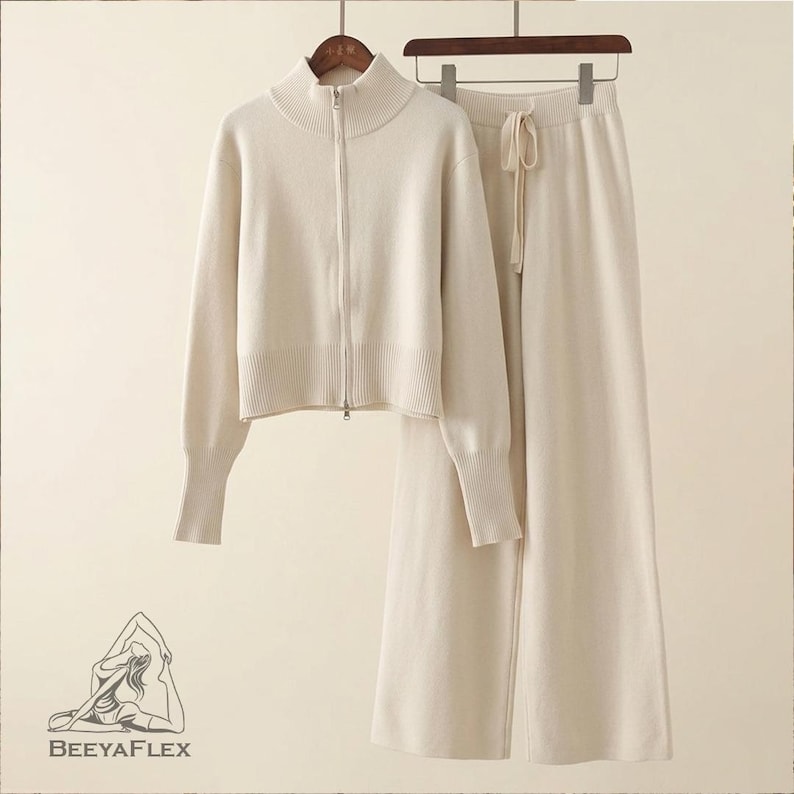 May include: A cream-colored knitwear set, including a zip-up jacket with a high collar and wide-leg pants with a drawstring waist. The set is displayed on a hanger against a neutral background. The jacket has long sleeves and ribbed cuffs. The pants are a solid color.