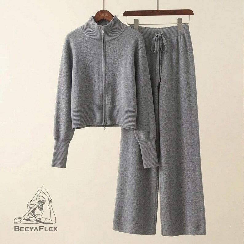 May include: A gray knit two-piece set, including a zip-up jacket with a high collar and matching wide-leg pants. The jacket has long sleeves and ribbed cuffs. The pants have a drawstring waist. The set is displayed on a hanger.