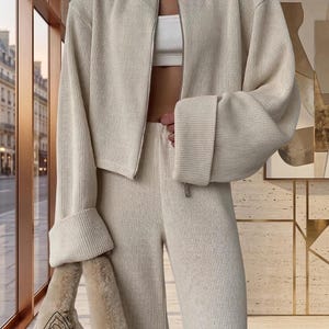 Puede incluir: Un conjunto de punto color crema, que incluye una chaqueta con cremallera y pu&ntilde;os enrollados y pantalones a juego. El conjunto se combina con un top corto blanco y un bolso de mano de piel sint&eacute;tica. La chaqueta tiene cierre de cremallera.