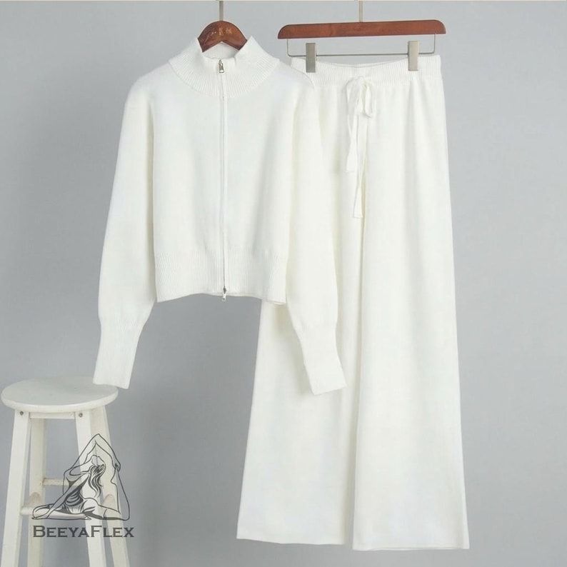May include: A white knit two-piece set, including a zip-up sweater with a high collar and wide-leg pants with a drawstring waist. The set is displayed on a hanger against a neutral background. The brand name "BEEAYAFLEX" is visible on a small stool in the foreground.