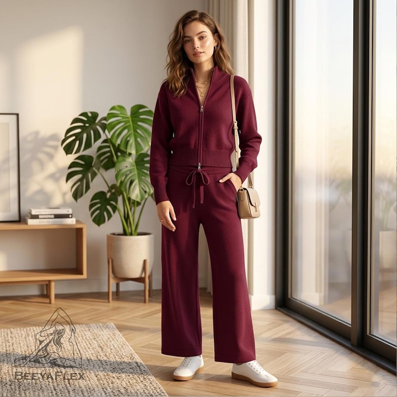 May include: A burgundy zip-up jacket and matching drawstring pants. The jacket has a high collar and the pants are wide-legged. The outfit is paired with white sneakers and a small beige crossbody bag. The outfit is made of a soft knit material.