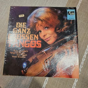May include: Vintage vinyl record album titled "Die Ganz Grossen Tangos" featuring Peter Kreuder's Tango-Orchester. The album cover shows a woman with a violin. The record is sealed in plastic and has a sticker indicating it is German.
