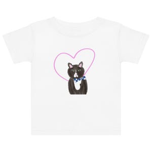 May include: White toddler t-shirt featuring a black cat illustration with a blue and white checkered bow tie, set against a pink heart outline. The cat has a grumpy expression. The shirt is made of a soft material.