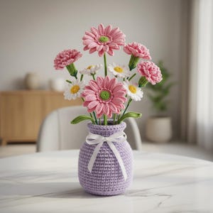Può includere: Un'arrangiamento floreale all'uncinetto fatto a mano in un vaso lavanda legato con un nastro bianco. Il bouquet presenta gerbere rosa, garofani e margherite bianche con centri gialli. Il vaso è su un tavolo di marmo bianco.