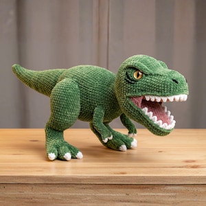 May include: A green crocheted Tyrannosaurus Rex toy with an open mouth, revealing white teeth and a pink interior. The dinosaur has yellow eyes and white accents on its feet. The toy is displayed on a wooden surface.