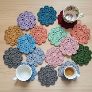 May include: A collection of handmade crochet coasters in various colors, including purple, teal, coral, and green. The coasters are arranged on a light wooden surface, with two teacups and a coffee cup resting on some of them. The coasters have a flower-like design.