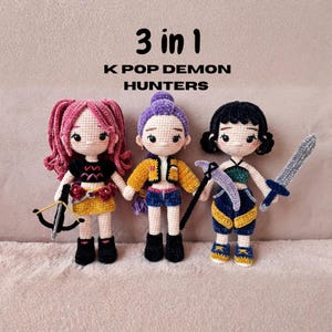Puede incluir: Tres muñecas de ganchillo hechas a mano con el texto "3 en 1 K POP DEMON HUNTERS". Cada muñeca tiene un atuendo y un arma únicos. Las muñecas tienen pelo rosa, morado y negro. Una muñeca sostiene un arco y flecha, otra una guadaña y la última una espada.