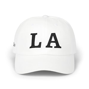 Op de afbeelding: Een witte baseball cap met de letters "LA" in vette zwarte letters op de voorkant. De cap heeft een gebogen klep en een knoop aan de bovenkant. Een eenvoudig, klassiek ontwerp geschikt voor dagelijks gebruik.