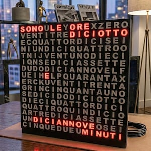 Può includere: Un orologio digitale nero mostra l'ora in lettere rosse. Le parole "VENTITREDICIOTTO" e "UNDICIANNOVEOSEI" sono evidenziate. L'orologio è su un tavolo di legno, con una vista della città sullo sfondo.