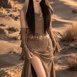 May include: A woman in a desert setting wears a long, flowing brown dress with a high slit. The dress features a hood and long sleeves with ruffled cuffs. A gold belt and headpiece add detail. Brown boots complete the look.