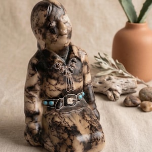 May include: A ceramic figurine of a seated figure with a detailed, patterned design in shades of brown and black. The figure wears a necklace and belt with turquoise accents. The overall aesthetic suggests a Native American theme.
