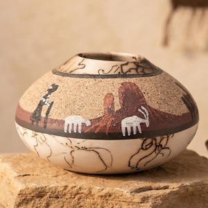 May include: Hand-painted ceramic pot with a rounded shape. The pot features a tan background with a brown band and a design depicting a desert landscape with white animals and a black figure. The rim has a dark pattern.