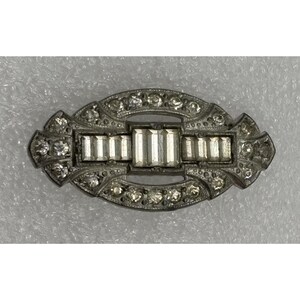 May include: An oval-shaped silver-tone metal brooch featuring a central horizontal row of rectangular baguette-cut clear stones flanked by smaller round faceted clear stones set within an intricate frame of additional round clear crystals.