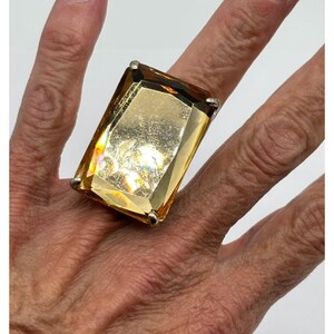 May include: A large rectangular ring featuring a faceted amber-colored gemstone set in a silver-toned metal prong setting. The stone has a polished surface with visible light reflections and covers a significant portion of the finger area.