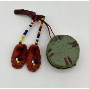 May include: A handcrafted Native American-style leather charm featuring two small red moccasins with beaded accents and a round, green leather pouch. The moccasins are adorned with blue, white, and yellow beads. The charm is attached to a brown leather strap.
