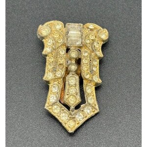 May include: A gold-tone metal dress clip featuring a symmetrical design adorned with numerous clear round rhinestones and a single rectangular baguette-cut rhinestone at the top center. The piece has an intricate, layered, and geometric structure.