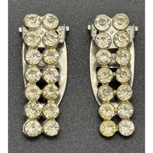 May include: A pair of silver-toned metal shoe clips featuring two vertical rows of clear, round, faceted rhinestones. Each clip has a rectangular base with a hinged mechanism on the back, designed to attach securely to footwear for added decoration.