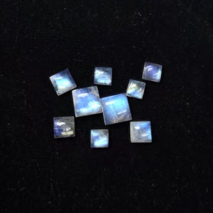 Puede incluir: Una colección de piedras preciosas de piedra lunar cuadradas e iridiscentes, exhibidas en una caja negra forrada de terciopelo. Las piedras exhiben un juego de colores azules y blancos, reflejando la luz. Las piedras preciosas están dispuestas en un patrón disperso.
