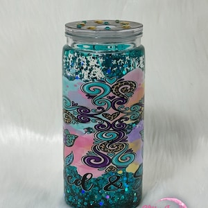 May include: A clear tumbler with a teal glitter base and lid. The tumbler features a colourful design with swirls, hearts, and leopard print accents. The words "feed & fit" are visible on the tumbler.