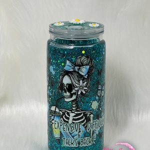 May include: A teal glitter tumbler with a skeleton graphic wearing sunglasses and a bow. The tumbler has the text "EXPENSIVE DIFFICULT TALKS BACK" and is topped with daisy accents.