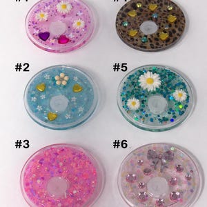 May include: Six round resin coasters with various decorative elements. Designs include glitter, flowers, hearts, and a bow. Each coaster is numbered for easy selection.