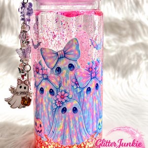 Puede incluir: Un vaso cilíndrico transparente lleno de líquido rosa y purpurina, con un gráfico de tres fantasmas con lazos y flores. La tapa está cubierta de cristales rosas. Un dije cuelga de la parte superior, mostrando un fantasma con una calabaza y el texto STAY SPOOKY.