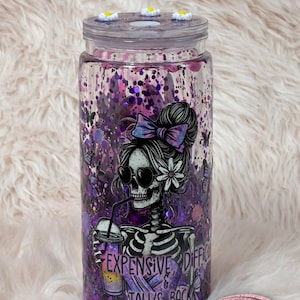 May include: A clear tumbler with a purple and pink glitter design. The tumbler features a skeleton graphic wearing sunglasses and a bow, holding a drink. The text on the tumbler reads "Expensive & Difficult Talks Back."
