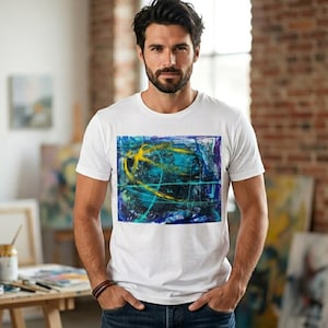 May include: White t-shirt with an abstract art print in blue, yellow, and purple. The design is dynamic and layered. The shirt is worn by a person in an art studio, with paintings and art supplies visible in the background.