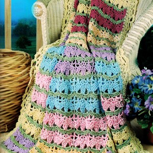 May include: A colorful, crocheted blanket draped over a wicker chair. The blanket features rows of pink, blue, green, and purple, with a yellow border. The chair is on a porch with a view of a landscape.