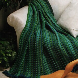 Pode incluir: Uma manta de crochê verde e azul drapeada sobre um sofá branco. A manta apresenta listras verticais com bolinhas texturizadas e uma franja azul escura. A paleta de cores inclui tons de verde e azul, criando uma estética aconchegante e convidativa.